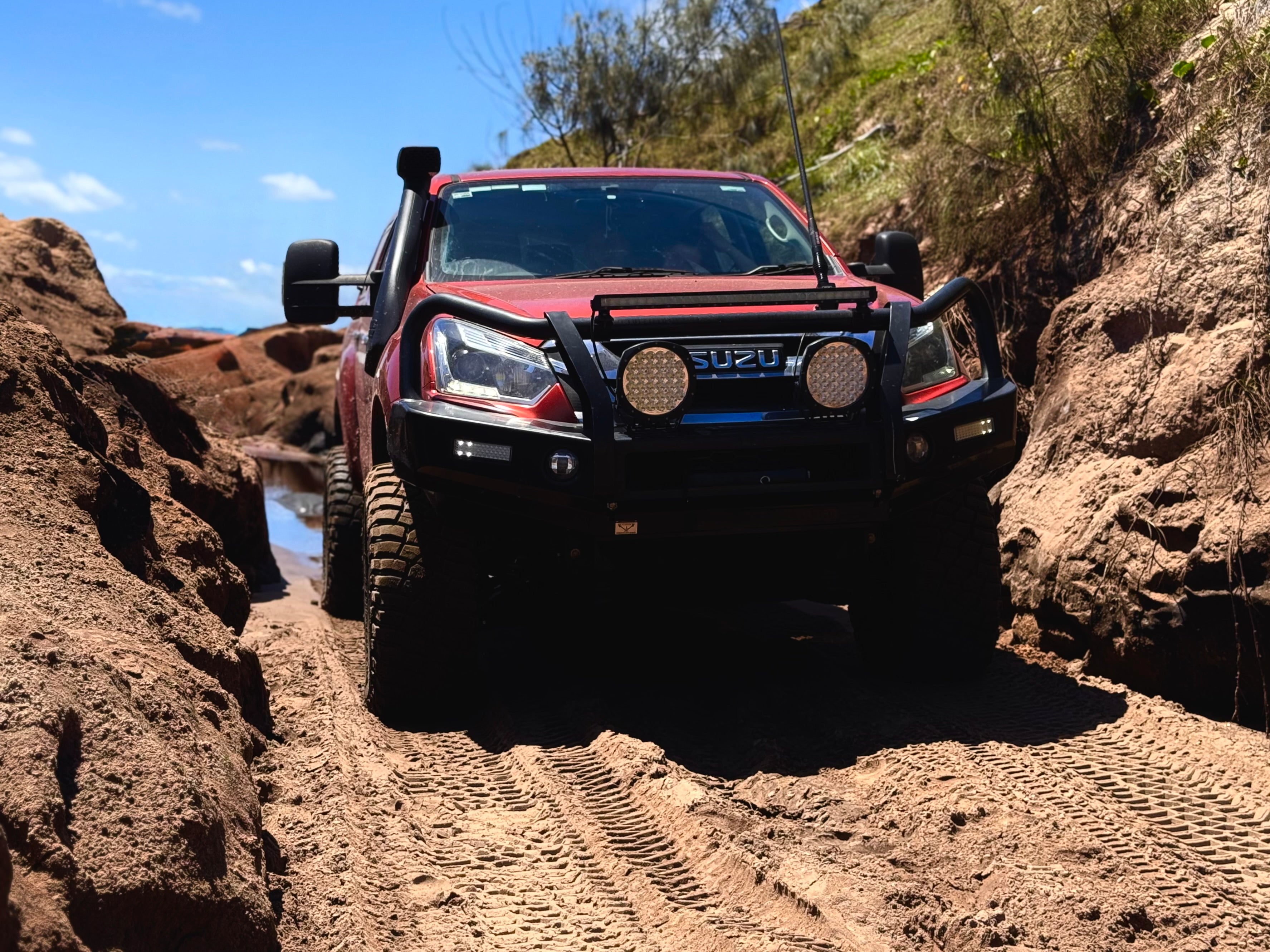 A red Isuzu D-Max 4x4 with a black bull bar, large off-road tires and a snorkel, navigates a sandy off-road track between two rugged rock formations.