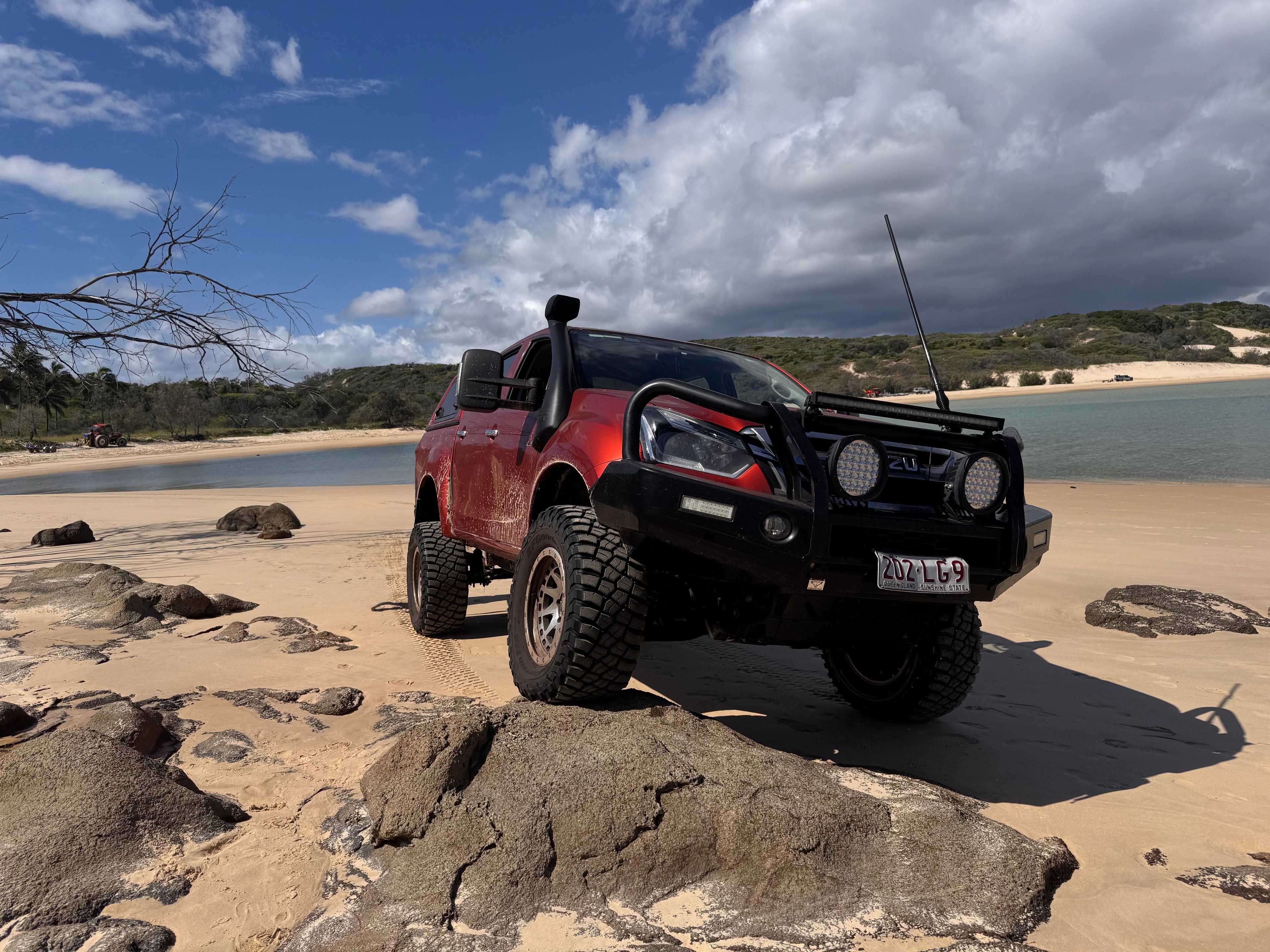 A red Isuzu D-Max is parked up on a rock in a rocky sandune with a lake in the background. The vehicle is heavily modified and sandy from off-roading on a rocky shorline.