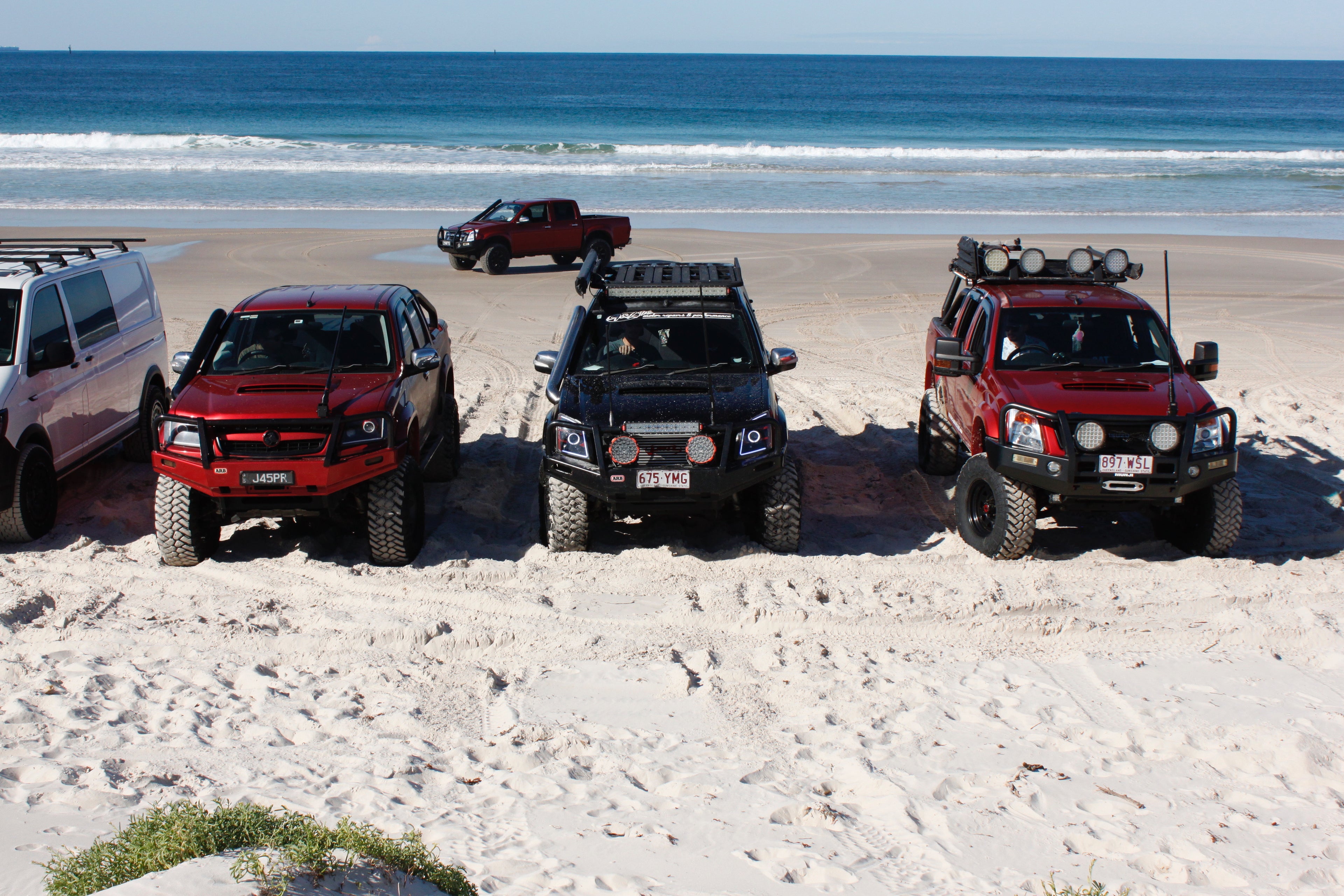 5 parked trucks at a beach, with sandy tires evident of off roading on sand.