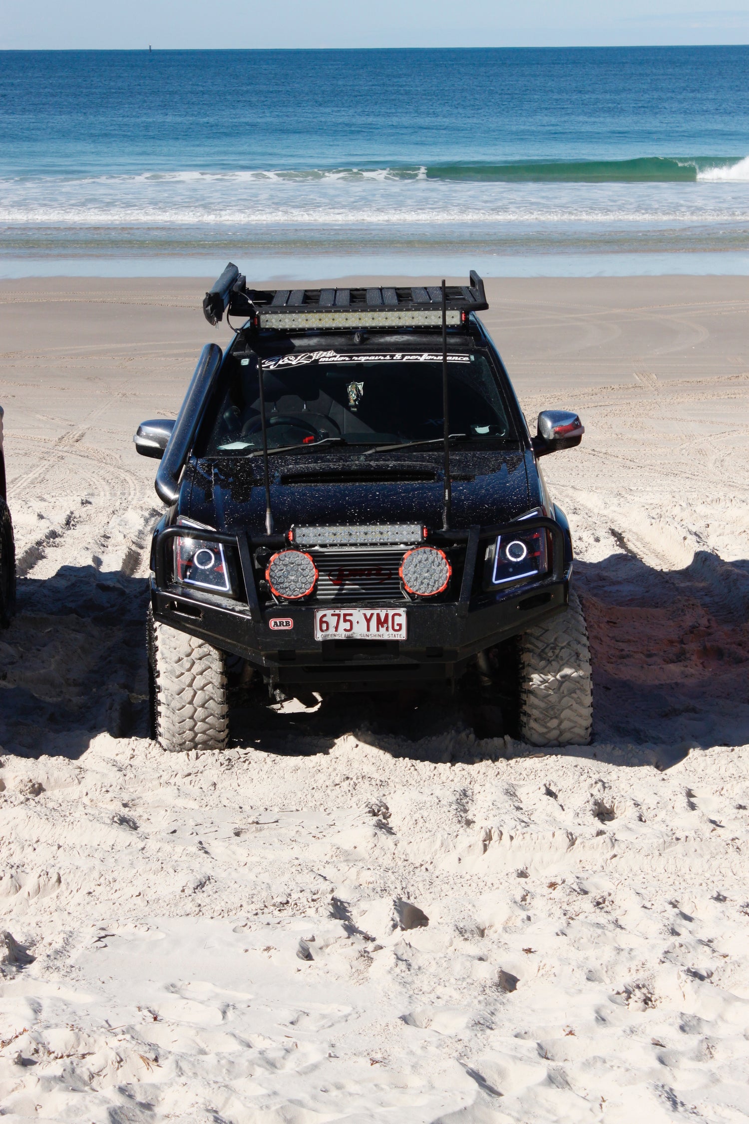 A black Holden Colorado Rc is parked on a sandy beach with calm waves in the background. The tires are sandy.